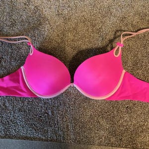 Puede incluir: Un sujetador rosa brillante con diseño push-up. El sujetador presenta un ribete blanco a lo largo de las copas y las correas, con correas rosas. El sujetador está diseñado con un cierre central y está hecho de un material liso, posiblemente similar al satén.