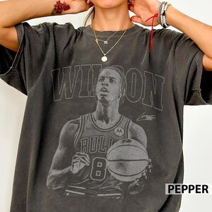以下が含まれることがあります： チャコールグレーの半袖Tシャツ。背番号8のユニフォームを着てバスケットボールを持つバスケットボール選手の、色あせたモノクロの大きなグラフィックプリントが施されており、上部には大きなブロック体でWILSONという文字がプリントされています。