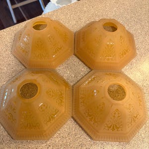 May include: Four vintage, amber-colored glass lamp shades. Each shade has an octagonal shape with a central hole and ornate, embossed floral patterns. The shades are arranged on a speckled countertop, showcasing their antique design.