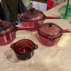 Puede incluir: Un juego de cuatro piezas de utensilios de cocina de vidrio rojo rubí vintage. El juego incluye dos cacerolas con tapas, una cacerola sin tapa y un plato redondo pequeño. Los utensilios de cocina son de vidrio transparente con asa.