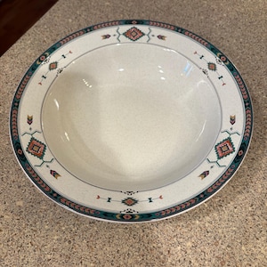 May include: A white ceramic mug with a black handle and the text "But First, Coffee" in a bold, black font. The mug is set against a blurred background, suggesting a cozy setting.