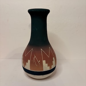 May include: A hand-painted ceramic vase with a dark teal neck and a bulbous body. The body features a design of white geometric shapes and a lightning bolt on a gradient of orange and brown. A dark blue band encircles the base.
