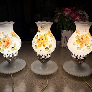 May include: Three vintage table lamps with white glass shades. Each shade is decorated with hand-painted yellow and orange roses and green leaves. The lamps have a white base and a decorative metal base. The lamps are lit, casting a warm glow.