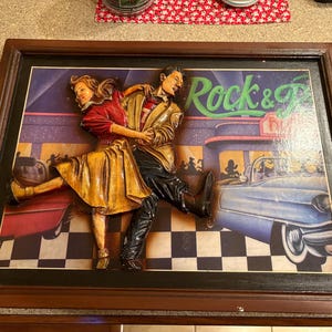 Pode incluir: Uma obra de arte 3D emoldurada que retrata um casal dançando em um cenário de restaurante retrô. O homem usa uma jaqueta bege e calças pretas, enquanto a mulher está com um top vermelho e uma saia amarela. As palavras "Rock & Roll" são visíveis.