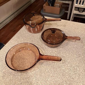 Puede incluir: Juego de utensilios de cocina ámbar de tres piezas. Incluye una cacerola con tapa, una olla pequeña con tapa y una sartén. Todas las piezas tienen asas y tapas a juego con pequeños pomos. El juego está hecho de vidrio transparente.