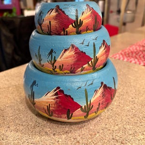 Puede incluir: Una pila de tres cuencos de madera pintados a mano. Cada cuenco es de color azul claro y presenta un diseño de paisaje desértico con montañas rojas, cactus y pájaros. Los cuencos están anidados.