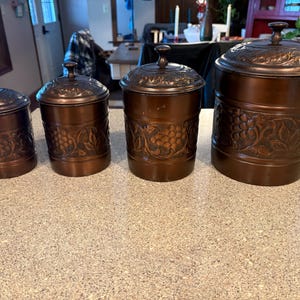 May include: Four copper-colored canisters with lids, arranged in a row. Each canister has a decorative embossed design, with the largest featuring a grape and leaf pattern. The canisters vary in size, with a matching lid on each.