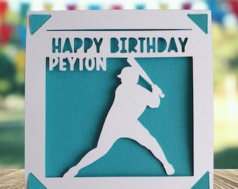 May include: A white baseball-themed birthday card with the words "HAPPY BIRTHDAY PEYTON" cut out at the top. A white silhouette of a baseball player is in the center, set against a teal background. The card is on a wooden surface.
