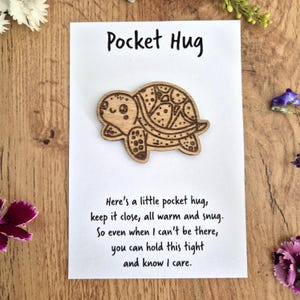 May include: A wooden turtle-shaped pocket hug on a white card with the text "Pocket Hug". The card also includes a poem about keeping the hug close. The turtle is light brown with etched details.