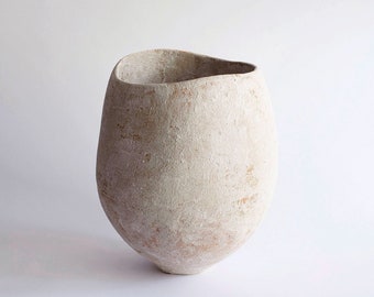 May include: A handcrafted, off-white ceramic vase with a textured surface and a unique, organic shape. The vase has a wide, irregular opening and a rounded base, perfect for displaying dried flowers or as a decorative accent.