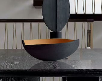 Puede incluir: Una mesa de comedor rectangular negra con una superficie texturizada y gruesas patas talladas. Un cuenco decorativo negro con interior dorado y una escultura abstracta negra están sobre la mesa. La mesa está rodeada de sillas blancas.