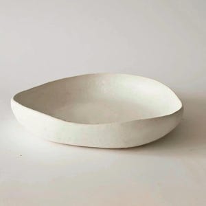 Pode incluir: Uma tigela de cerâmica branca suja feita à mão, com um acabamento liso e fosco. A tigela tem uma forma orgânica e assimétrica e uma profundidade rasa, adequada para servir ou exibir. A cor neutra e o design minimalista tornam-na uma peça versátil.