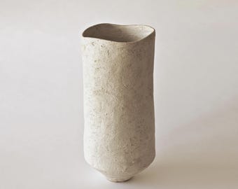 May include: A tall, off-white ceramic vase with a textured surface. The vase has a slightly irregular, organic shape with a flared opening and a rounded base. The neutral color and minimalist design make it suitable for various decor styles.