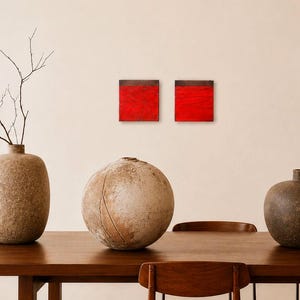 Puede incluir: Una mesa de comedor de madera con tres jarrones decorativos y dos pinturas cuadradas rojas. Los jarrones son texturizados y en tonos marrones. Una rama con hojas está en un jarrón. Una lámpara moderna cuelga encima.