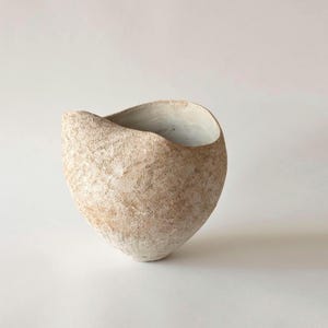 May include: A textured, beige ceramic bowl with a unique, asymmetrical shape. The interior is painted white, contrasting with the exterior's rough, earthy appearance. The bowl's design suggests a handcrafted aesthetic, suitable for decorative or functional use.