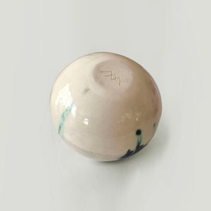 May include: A small, white ceramic bowl with a green and black glaze. The bowl has a round shape and a smooth, glossy finish. The bottom of the bowl is marked with a signature.
