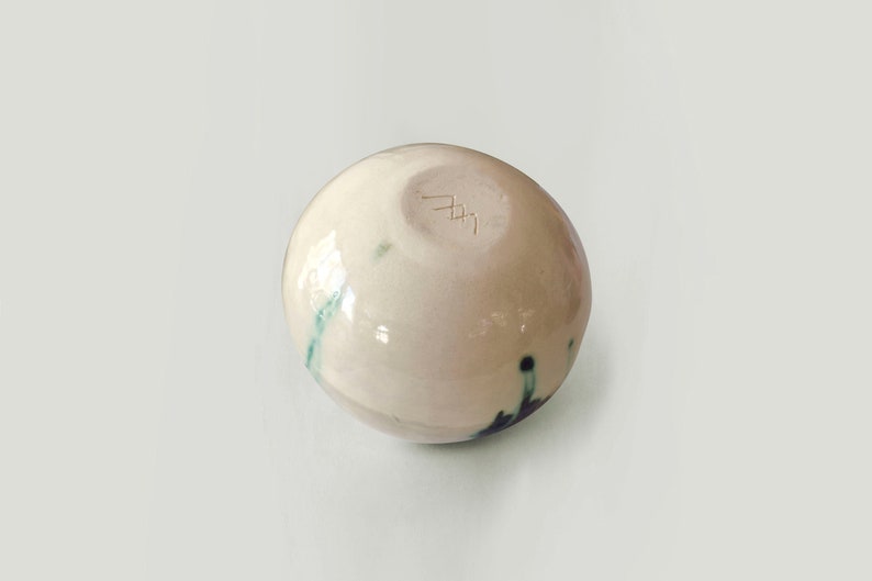 May include: A small, white ceramic bowl with a green and black glaze. The bowl has a round shape and a smooth, glossy finish. The bottom of the bowl is marked with a signature.