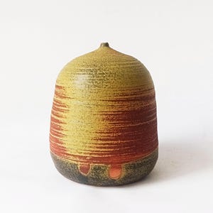 Può includere: Un vaso in ceramica fatto a mano con una forma arrotondata e strutturata. Il vaso presenta fasce orizzontali di tonalità giallo e rosso-arancio, con una base verde scuro e marrone. La parte superiore ha una piccola punta. Un pezzo unico per l'arredamento.