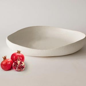 May include: A large, off-white, oval-shaped bowl with a textured surface. Three pomegranates, two whole and one halved, are arranged in front of the bowl. The pomegranates are a vibrant red with visible seeds.