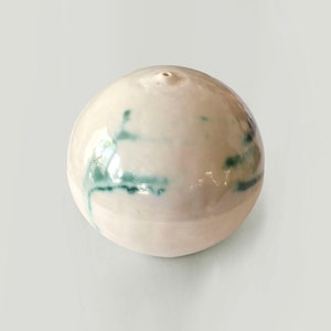 May include: A white ceramic sphere with a small hole at the top and green streaks throughout the surface.