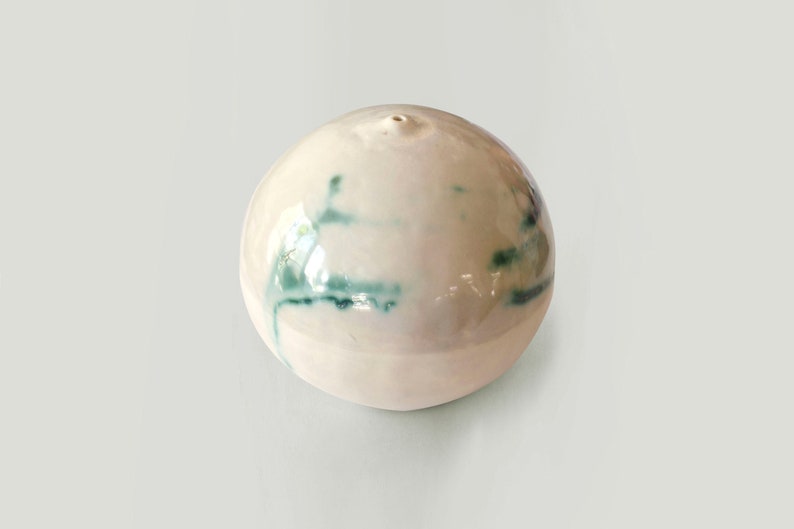 May include: A white ceramic sphere with a small hole at the top and green streaks throughout the surface.