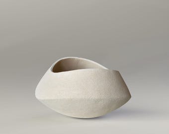 May include: A textured, off-white ceramic bowl with an asymmetrical, organic shape. The bowl has a wide opening and a faceted exterior, creating a modern, sculptural aesthetic. The neutral color and unique design make it a versatile decorative piece.