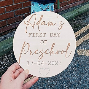 May include: A round wooden sign with the text "Adam's First Day of Preschool 17-04-2023" in gold lettering. A small heart is below the date.