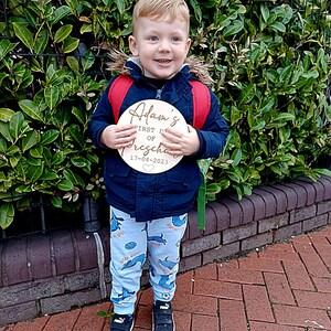 May include: A young boy wearing a blue jacket and blue and white patterned pants holds a wooden sign that says "Adam's First Day of Preschool 17-04-2023".