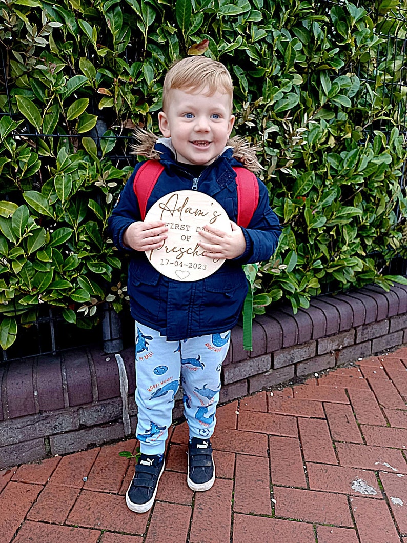 May include: A young boy wearing a blue jacket and blue and white patterned pants holds a wooden sign that says "Adam's First Day of Preschool 17-04-2023".