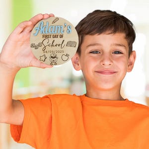 May include: A child is holding a round wooden sign that reads "Adam's First Day of School 04/09/2025". The sign is decorated with school-themed illustrations, including a pencil, a book, a star, a graduation cap, and an apple.