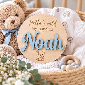 Puede incluir: Un letrero redondo de madera para anuncio de nacimiento con el texto grabado Hello World my name is y el nombre Noah en letras cursivas azul claro en relieve, junto a un pequeño icono de oso grabado en madera clara.