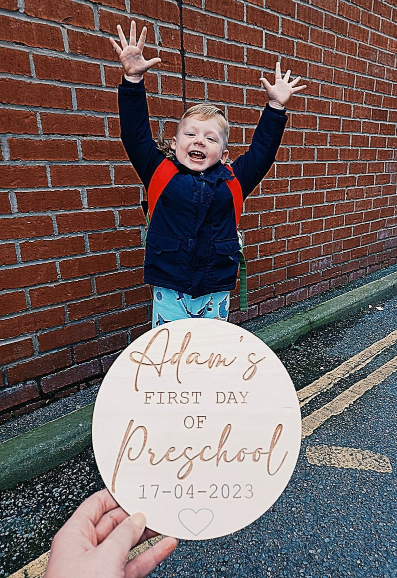 May include: A round wooden sign with the text "Adam's First Day of Preschool 17-04-2023" in gold lettering. A small heart is below the date.