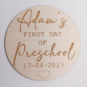 May include: A round wooden sign with the text "Adam's First Day of Preschool 17-04-2023" and a small heart.