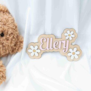 May include: A wooden name sign with the name "Ellery" in pink letters. The sign is decorated with white daisies with yellow centres.