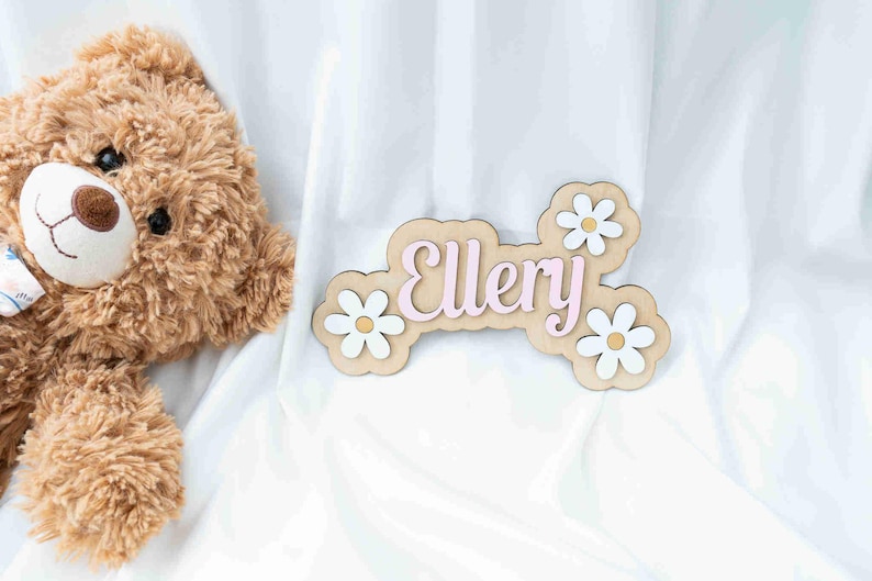 May include: A wooden name sign with the name "Ellery" in pink letters. The sign is decorated with white daisies with yellow centres.