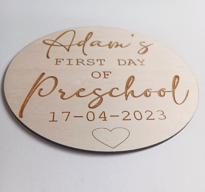 May include: A round wooden sign with the text "Adam's First Day of Preschool 17-04-2023" and a small heart.