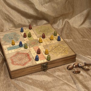 May include: A square wooden board game with a hinged metal latch, featuring a printed grid and four decorative mandala-style patterns in the corners. Small, cone-shaped game pieces in blue, yellow, green, and red are placed on the board.