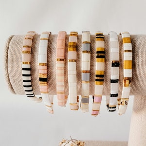 May include: A set of ten colorful beaded bracelets with gold accents. The bracelets are arranged in a row on a beige fabric stand.