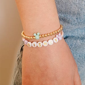 May include: A close-up of a person's wrist wearing two bracelets. The top bracelet is gold with a light green Mickey Mouse charm. The bottom bracelet is white with gold lettering spelling out "DISNEY".