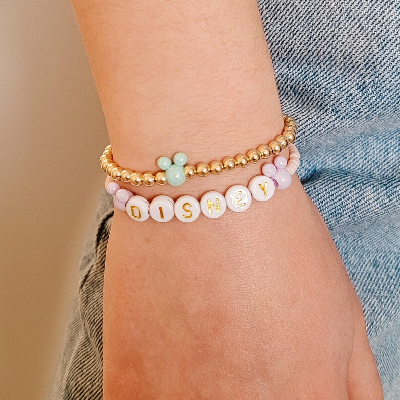 May include: A close-up of a person's wrist wearing two bracelets. The top bracelet is gold with a light green Mickey Mouse charm. The bottom bracelet is white with gold lettering spelling out "DISNEY".