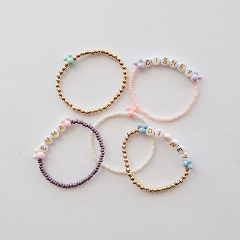 May include: Four beaded bracelets with different colored beads and charms. The bracelets spell out the names "Emma", "Disney", and "Disney".