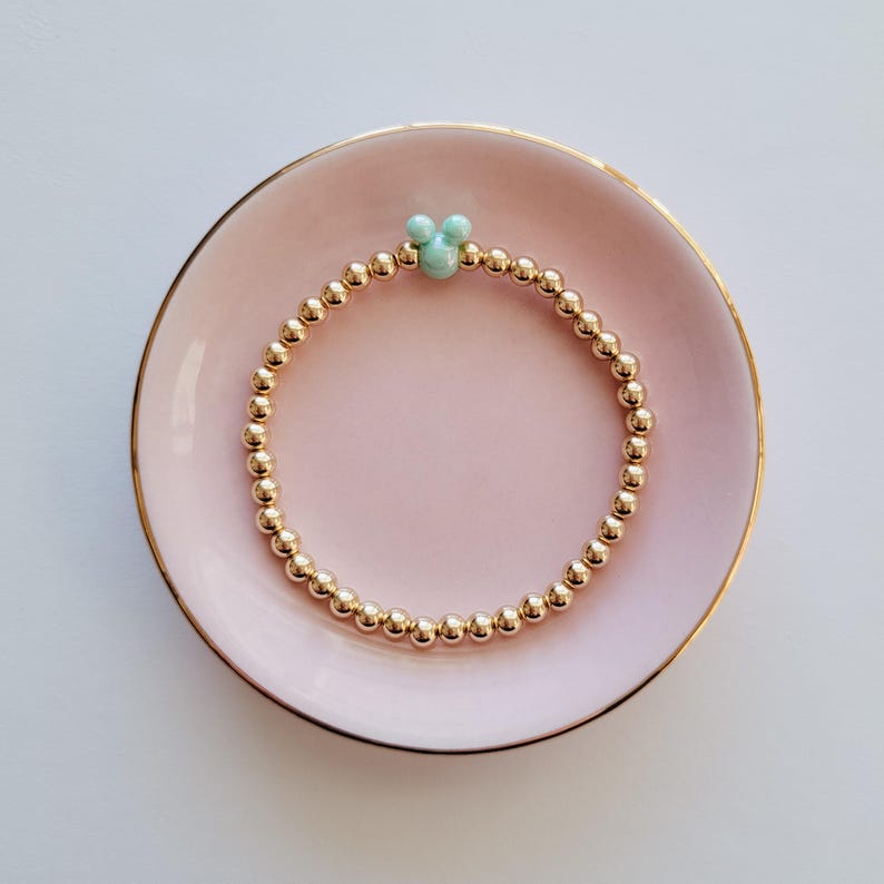 May include: A gold beaded bracelet with a light blue Mickey Mouse charm. The bracelet is on a pink dish.