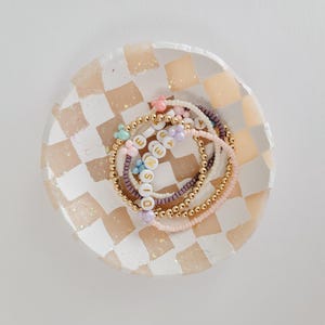 May include: A set of colorful beaded bracelets with the word "DISNEY" spelled out in gold letters. The bracelets are arranged in a checkered dish with a pink and white design.
