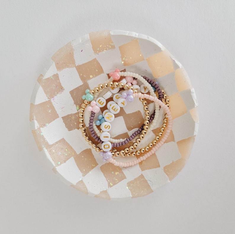 May include: A set of colorful beaded bracelets with the word "DISNEY" spelled out in gold letters. The bracelets are arranged in a checkered dish with a pink and white design.