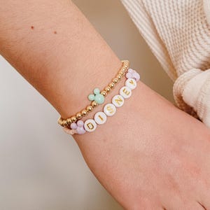 May include: A gold beaded bracelet with a light green Mickey Mouse charm and a white beaded bracelet with the word "DISNEY" spelled out in gold letters.