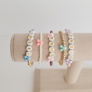 May include: Five beaded bracelets with gold beads and letter beads spelling out the names "DISNEY", "EMMA", and "DISNEY". The bracelets are displayed on a beige fabric stand.