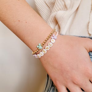 May include: A gold beaded bracelet with a white bead charm that spells out "DISNEY" and a small green Mickey Mouse charm.