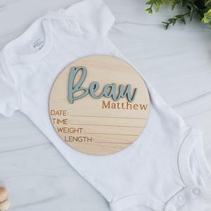 Round wooden baby birth announcement plaque placed on a white infant onesie. The plaque features the name 'Beau Matthew' with 'Beau' in raised blue 3D letters and 'Matthew' engraved below. It includes engraved sections labeled Date, Time, Weight, and Length. The setup is styled with greenery and wooden baby toys on a light marble background.