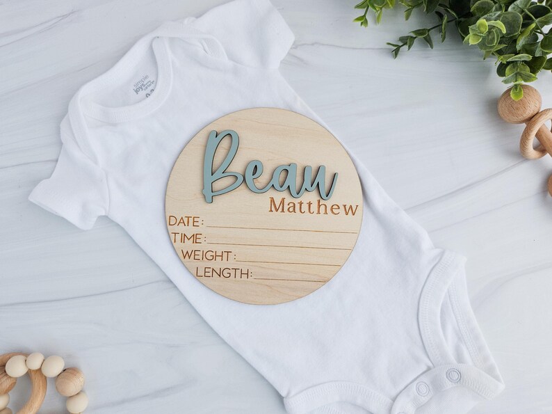Round wooden baby birth announcement plaque placed on a white infant onesie. The plaque features the name 'Beau Matthew' with 'Beau' in raised blue 3D letters and 'Matthew' engraved below. It includes engraved sections labeled Date, Time, Weight, and Length. The setup is styled with greenery and wooden baby toys on a light marble background.