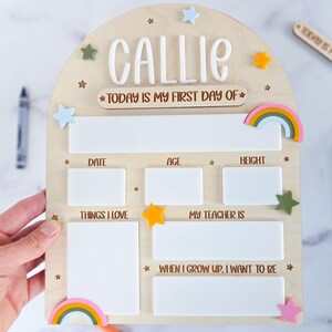 May include: An arched wooden sign with the name CALLIE in white 3D letters and the text TODAY IS MY FIRST DAY OF. It features white rectangular spaces for writing details like date, age, height, things I love, teacher, and future goals, decorated with small colorful stars and rainbows.